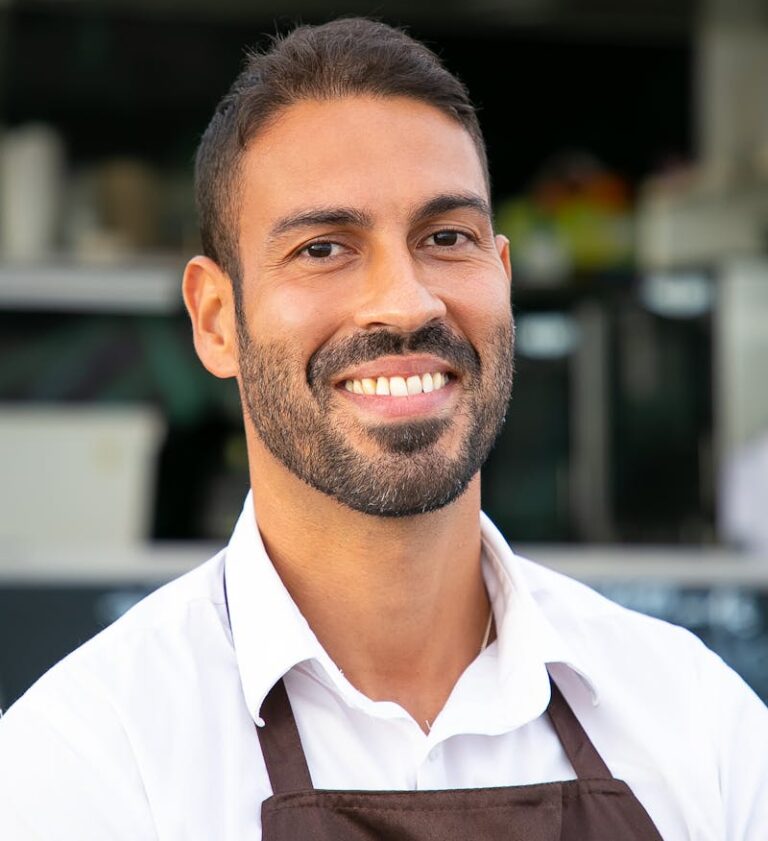 Cheerful ethnic waiter in apron standing against street cafe